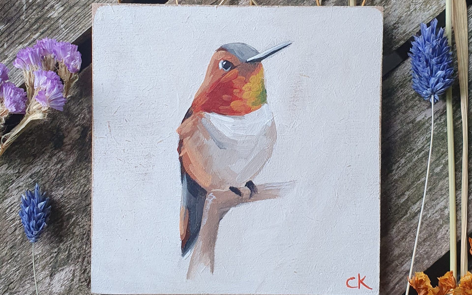 Kolibrie op een takje. Kolibrie met rood oranje en groene kleuren bij de hals. Crème kleurig buikje en lichte achtergrond. 10 x 10 cm paneeltje met goudkleurig haakje, klaar om op te hangen.