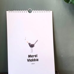 An A4 size calendar standing on wood with a green wall behind it. It has wires and a hook to hang. White front page with a little blackbird illustrated on it. Text says Merel Vlekkie 2023.