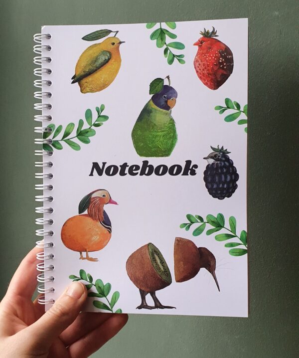 Notitieboekje A5 formaat, vastgehouden door een hand tegen een achtergrond van een groene muur. Witte voorkant met verschillende kleurige vogels in de vorm van fruit. Tekst in donkerblauw: Notebook.